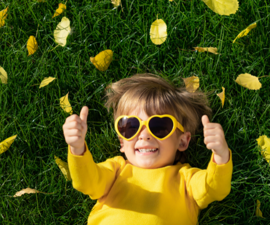 Toddler Laying in Green Grass with Leaves giving thumbs up.